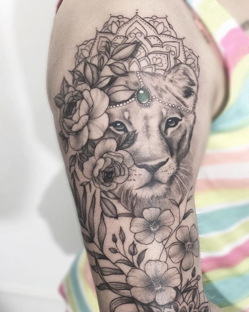 w819-35 Tatuagem no braço de leão rodeado por flores e folhas, com detalhes ornamentais e uma pedra verde na testa.