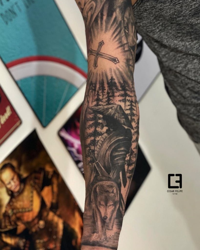w819-39 Braço tatuado com imagens de cruz, cavaleiro medieval e lobo em meio a árvores, em frente a quadros na parede.