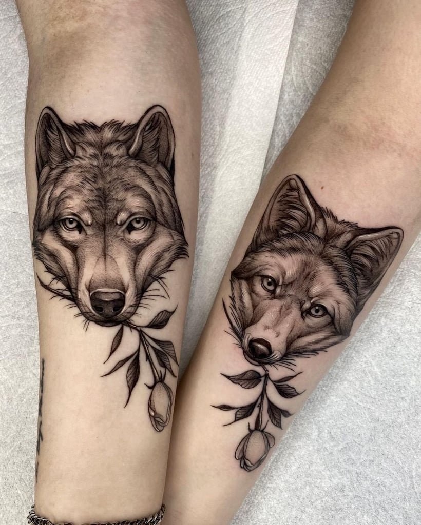 w820-15 Tatuagens de lobos detalhados em dois braços, cada um segurando um ramo com folhas e flores.