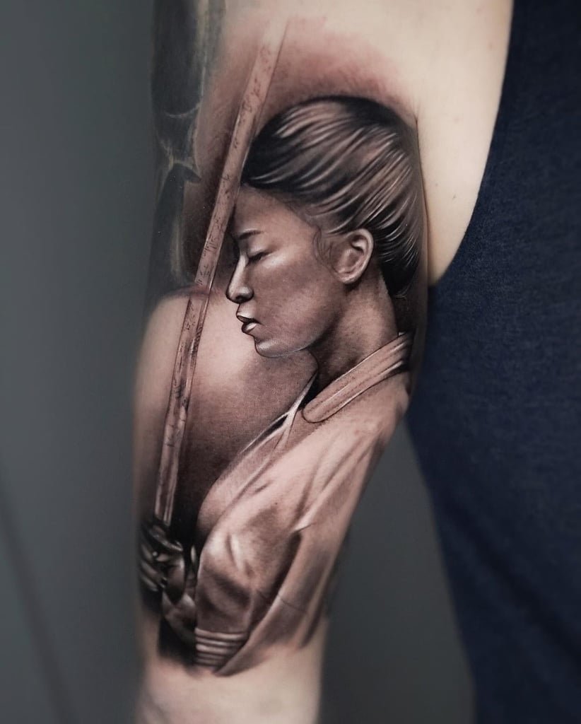 w822-2 Tatuagem realista de uma pessoa segurando uma espada, com olhos fechados e cabelo preso, no braço de alguém.