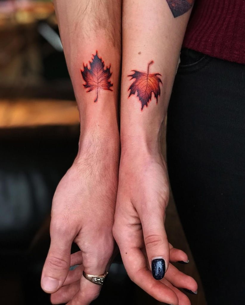 w822-8 Dois antebraços com tatuagens de folhas vermelhas, mãos entrelaçadas, uma com esmalte escuro.