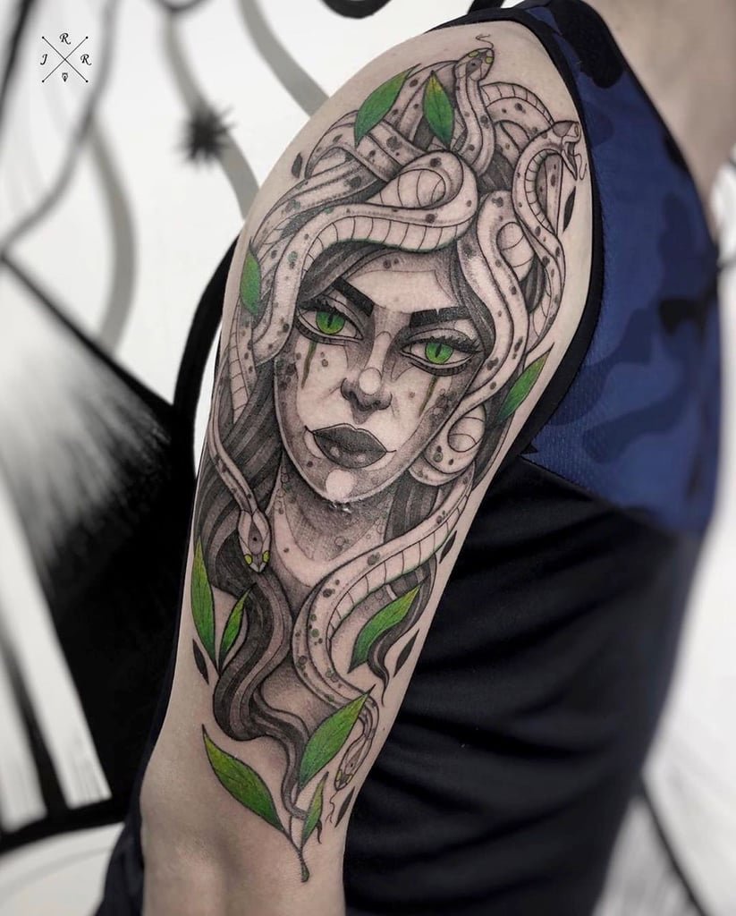 w823-2 Tatuagem de figura mitológica com cobras e folhas verdes no braço de uma pessoa usando camiseta azul.