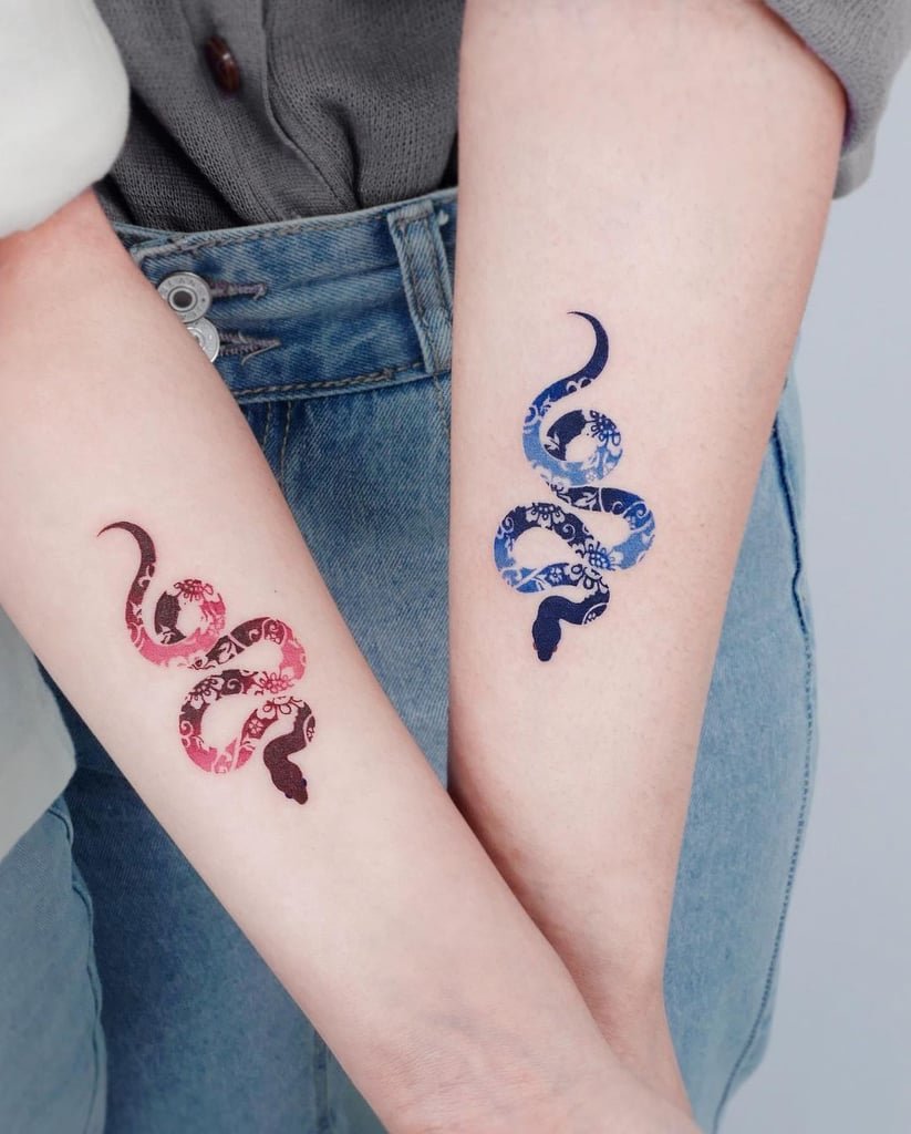 w823-6 Dois braços com tatuagens de cobras coloridas, uma em rosa e outra em azul, com detalhes florais.