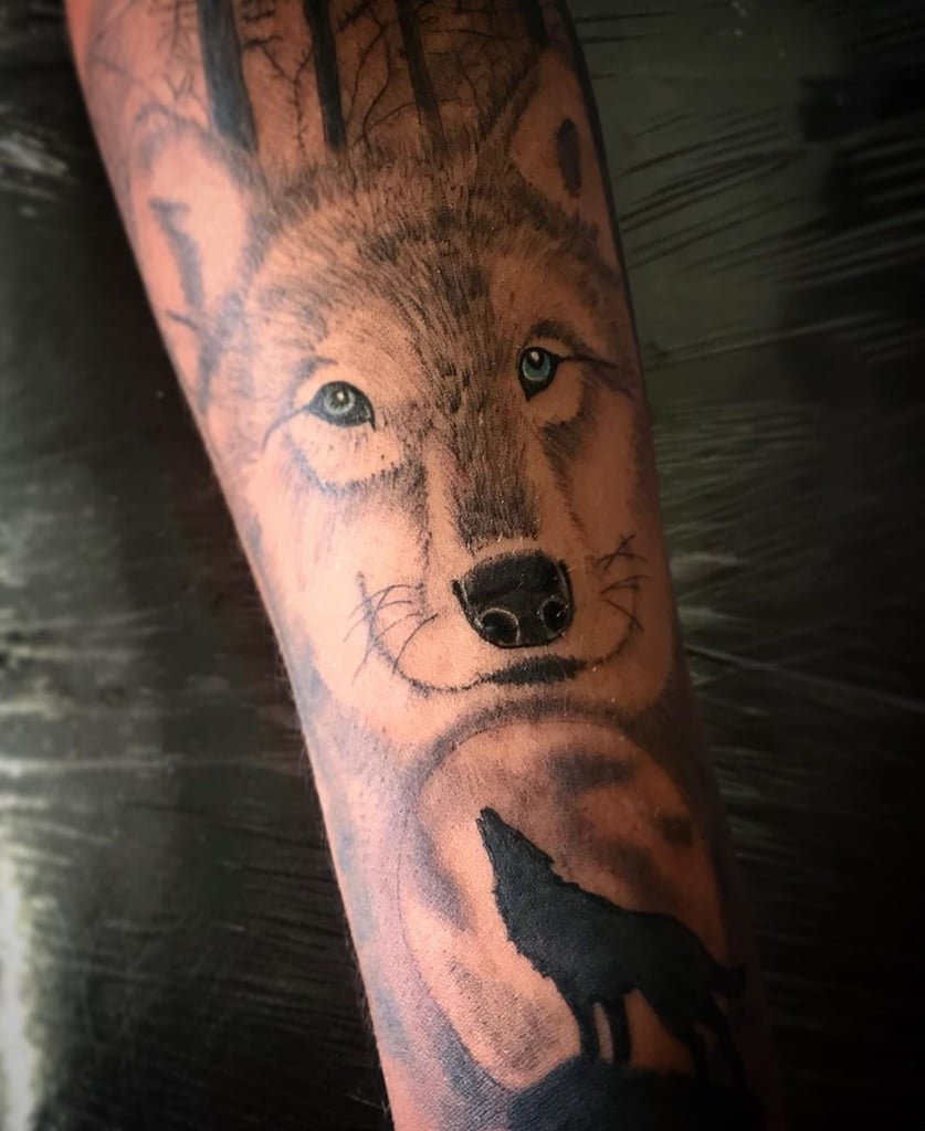 w837 Tatuagem de lobo realista com silhueta de lobo uivando sob a lua cheia e árvores ao fundo.
