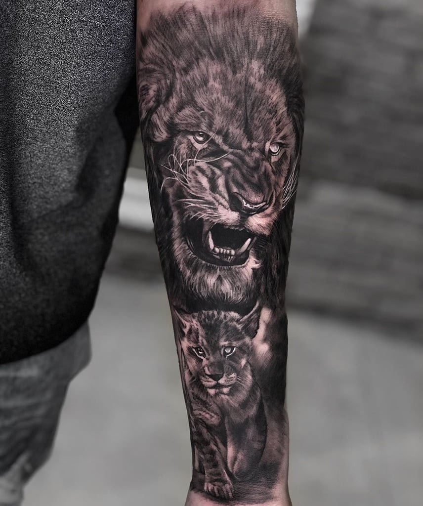 w856 Braço tatuado com leões realistas, um rugindo e outro filhote, em tons de preto e cinza.