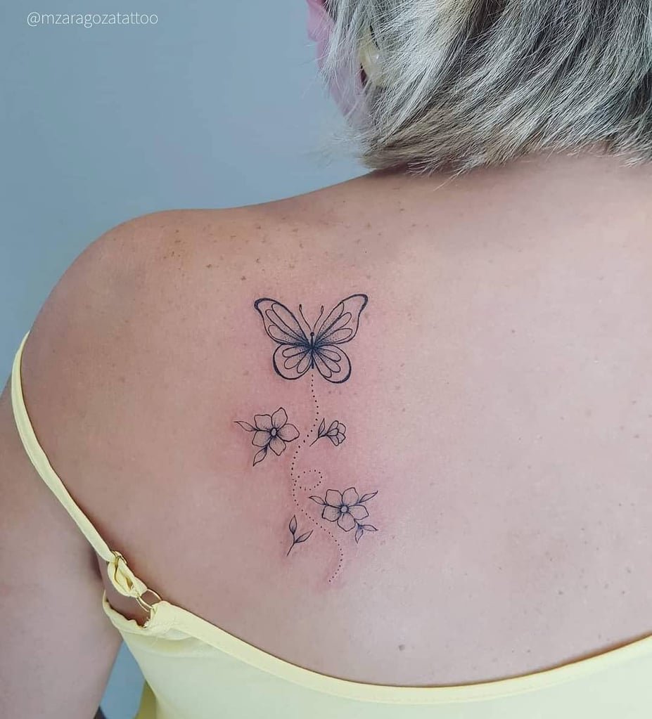 w927-1 Uma tatuagem de borboleta e flores delicadas no ombro de uma pessoa com cabelo loiro curto, usando blusa amarela.