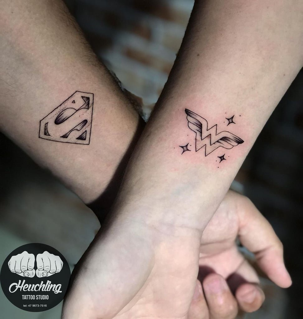 w973 Duas tatuagens de logotipos em antebraços; uma com um "S" estilizado, outra com um "W" com asas e estrelas.