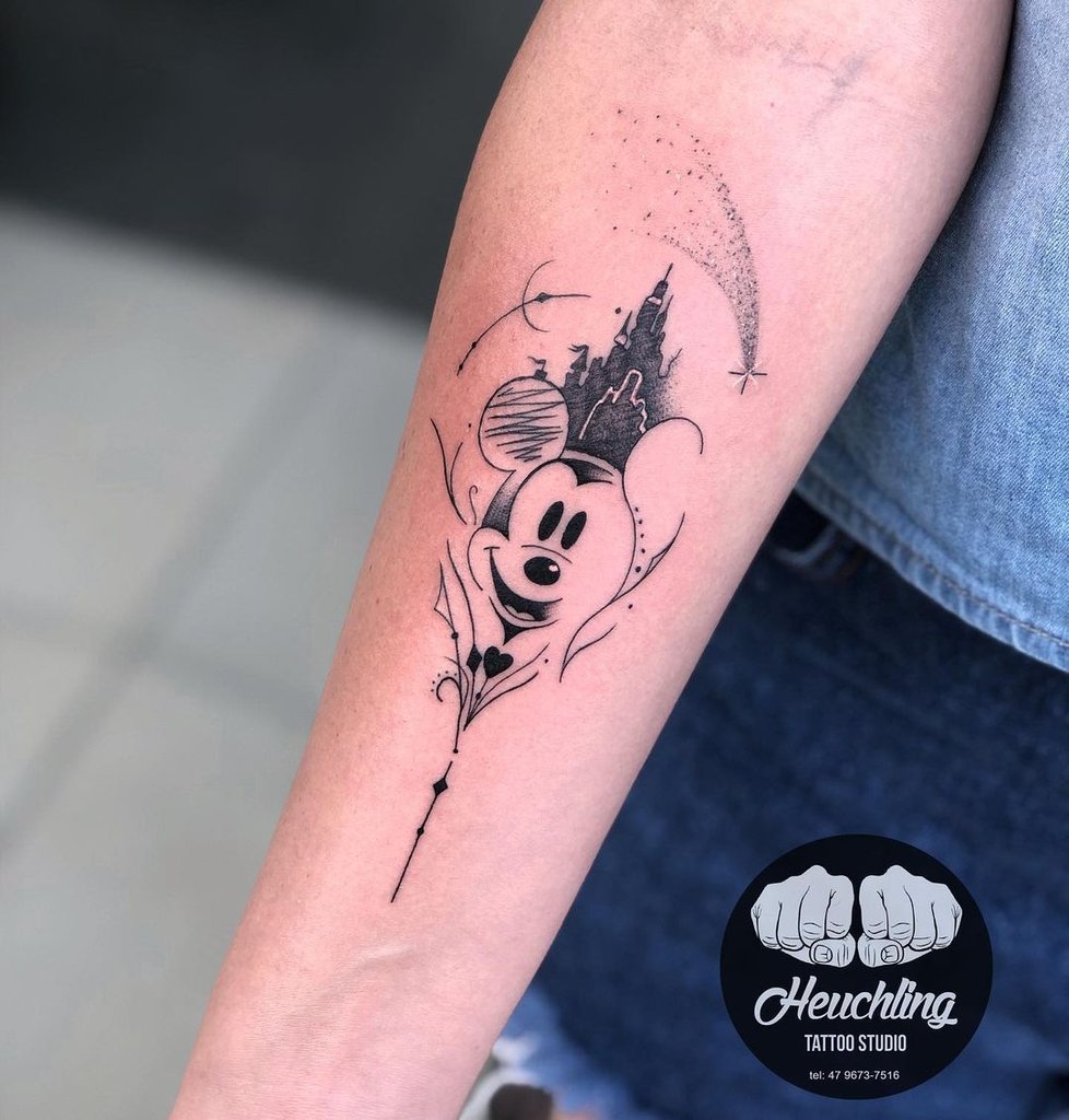 w978 Um braço com uma tatuagem em preto e branco de um personagem animado famoso, com elementos decorativos e um castelo.