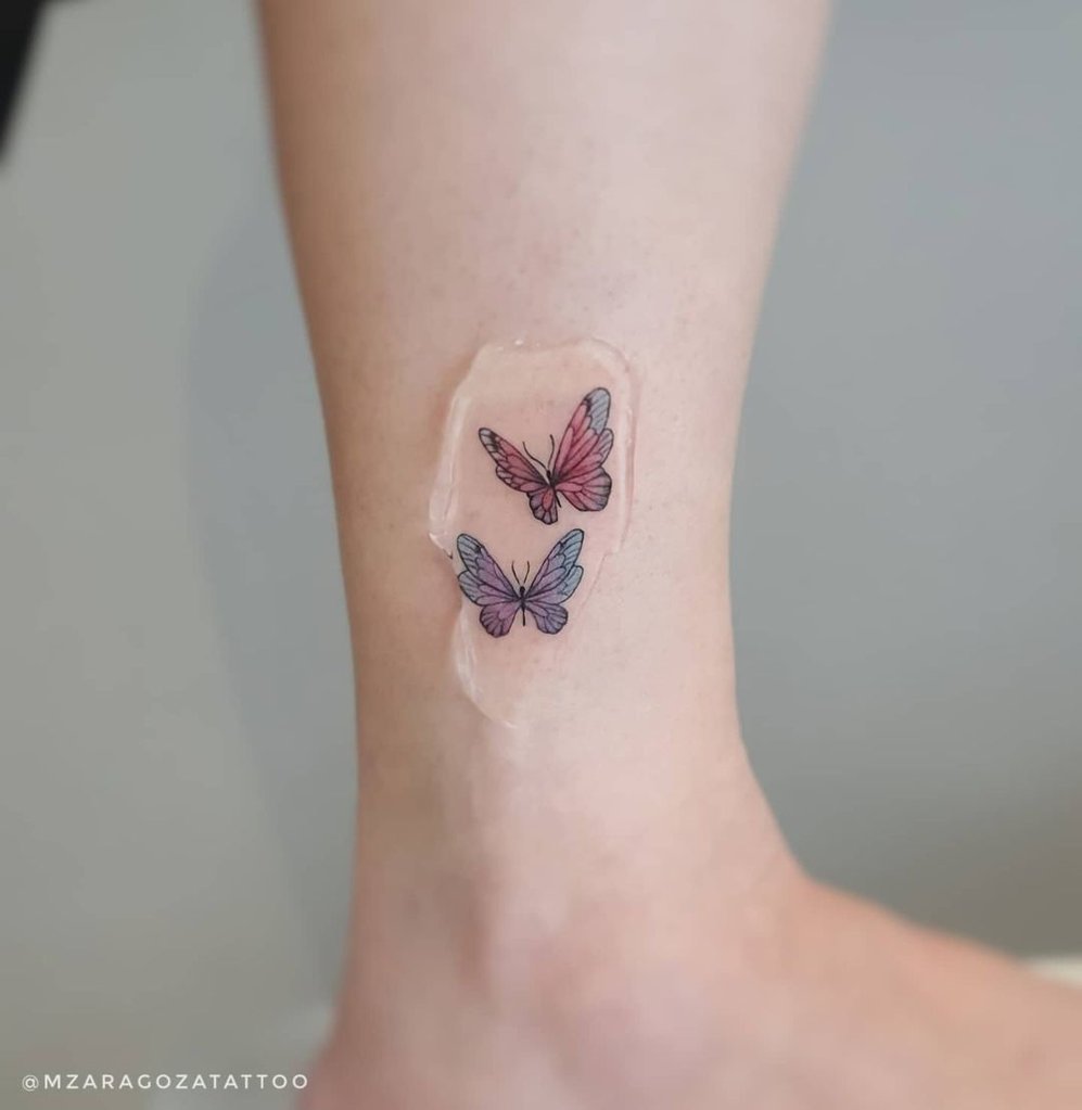 w997-1 Duas tatuagens de borboletas coloridas no tornozelo, cobertas por uma película protetora transparente.