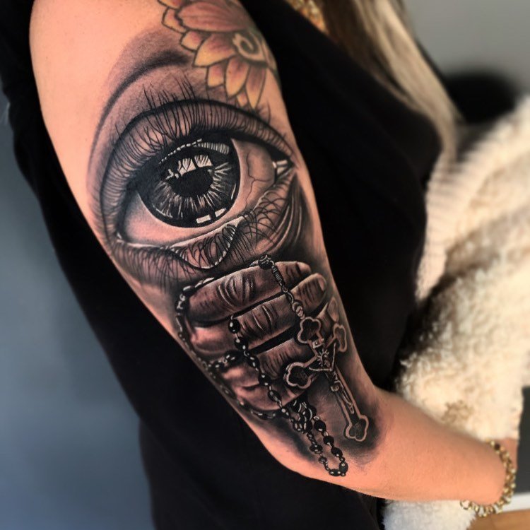 w9999-7 Tatuagem no braço de um olho grande e uma mão segurando um rosário detalhado, com flores no fundo.