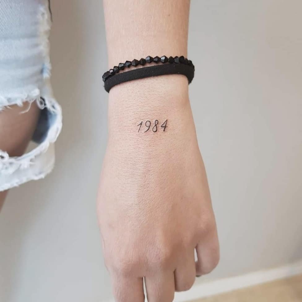 w9999-84 Pessoa com tatuagem "1984" no pulso e pulseira preta.