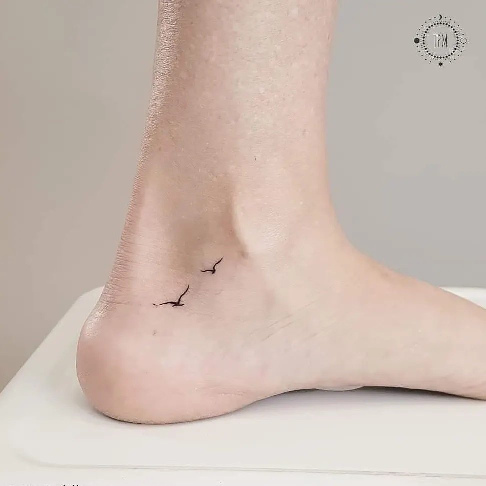 w9999-86 Pessoa com tatuagem de pássaros voando no tornozelo, fotografada de perfil, sobre superfície branca.
