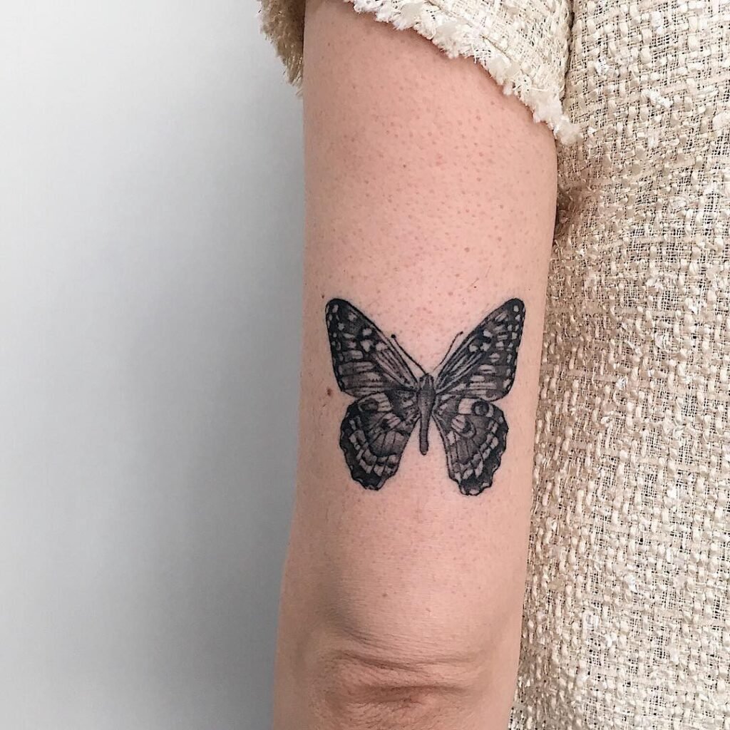 Tatuagem de borboleta em preto e branco no braço de uma pessoa com blusa de tecido texturizado.