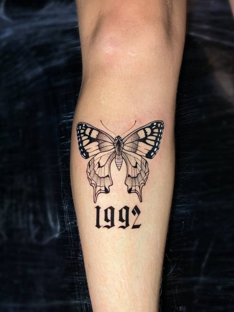 abaixo-joelho-borboleta Tatuagem de borboleta na canela de uma pessoa de pele clara com sombreamento, traços finos e grossos e o número 1992 tatuado abaixo.