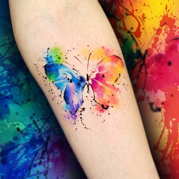 aquarela-azul-verde-rosa-roxo-borboleta Tatuagem estilo aquarela em tons de azul, verde, rosa e roxo no braço de uma mulher de pele clara.