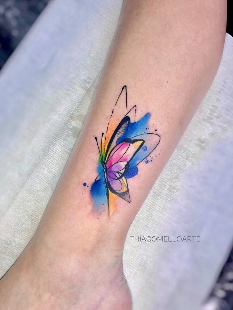 aquarela-estilo-borboleta Tatuagem de borboleta com traços finos e grossos e desenho um pouco mais minimalista, mas com efeito aquarela nas cores azul, rosa e amarelo. Foi feita no braço de uma mulher de pele clara.