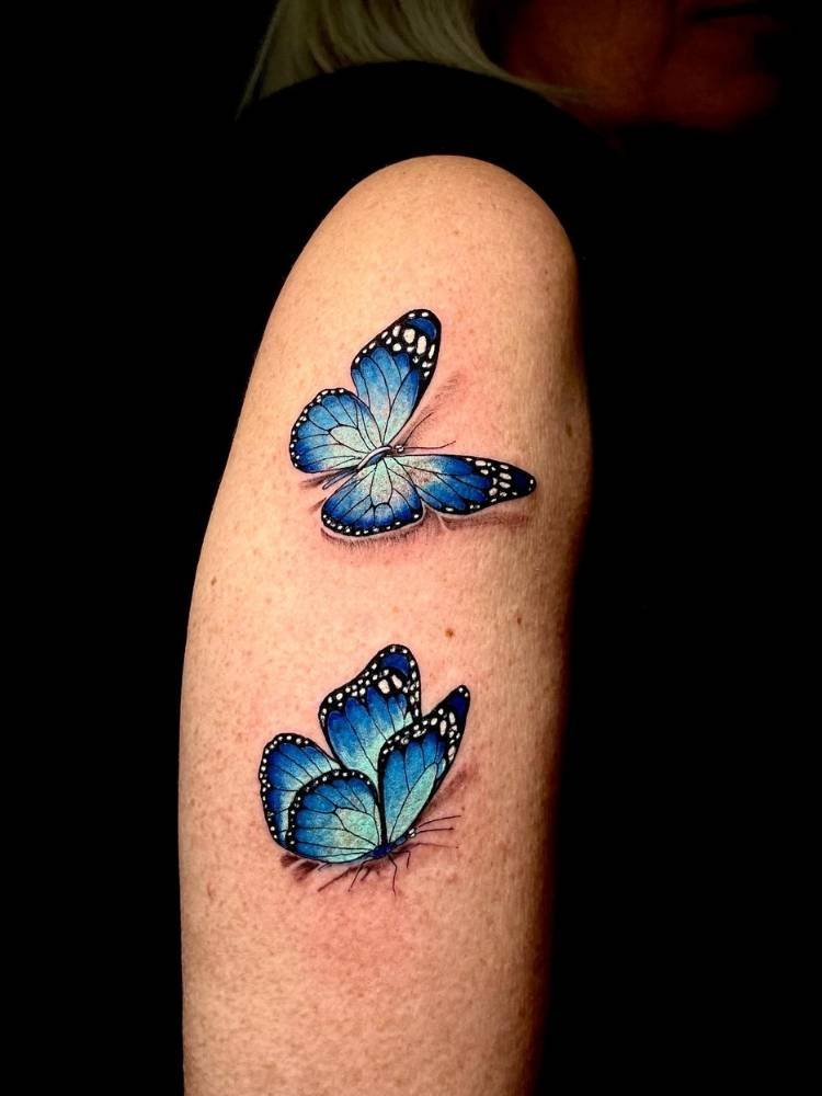 azul-sombreamento-borboleta Braço de uma mulher de pele clara com duas borboletas delicadas azuis tatuadas com um leve sombreamento.