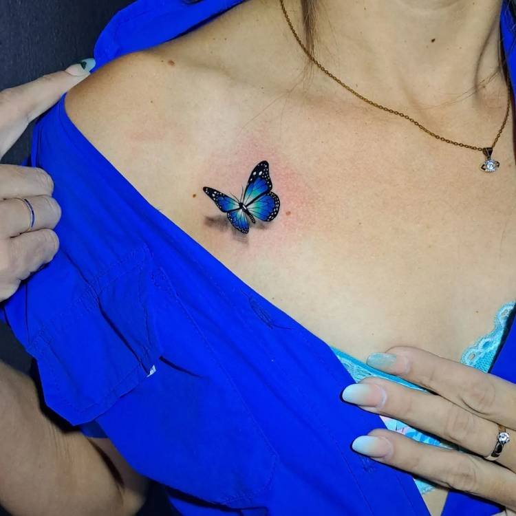 azul-sombreamento-ombro-borboleta Pequena tatuagem de borboleta azul com sombreamento que dá efeito 3D de pouso do inseto no ombro de uma mulher de pele clara vestida com uma roupa azul.