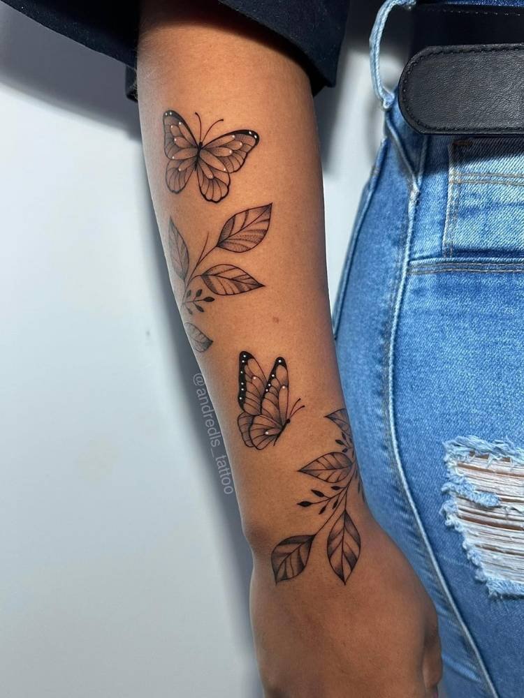 borboleta-com-ramos Tatuagem de borboleta com ramos em traço fino no braço de uma mulher com pele morena, usando blusa preta e calça jeans.