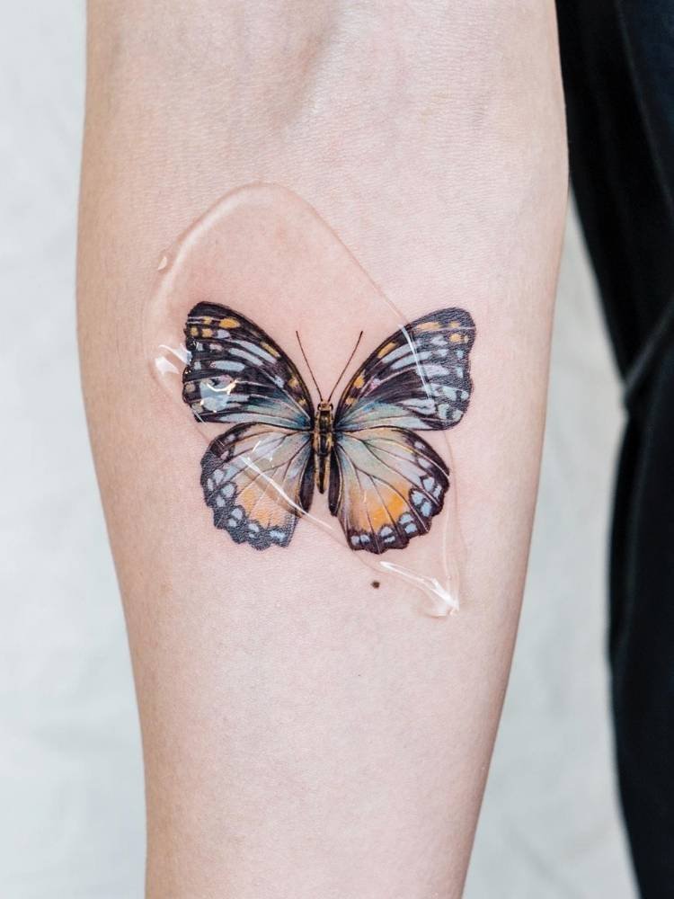 colorida-azul-amarelo-braco Tatuagem realista e cheia de detalhes de borboleta em tons de azul claro e amarelo no braço de uma pessoa de pele clara.