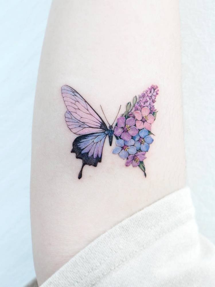 colorida-azul-roxo-rosa-flor Tatuagem de borboleta azul, rosa e lilás cheia de detalhes. Feita no braço de uma mulher de pele clara.