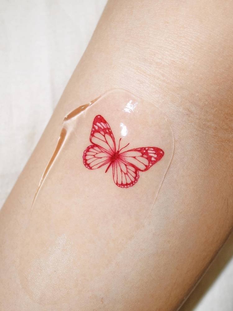 colorida-traco-vermelho-borboleta Tatuagem pequena de borboleta com traços finos e um pouco mais grossos em vermelho no braço de uma mulher de pele clara.