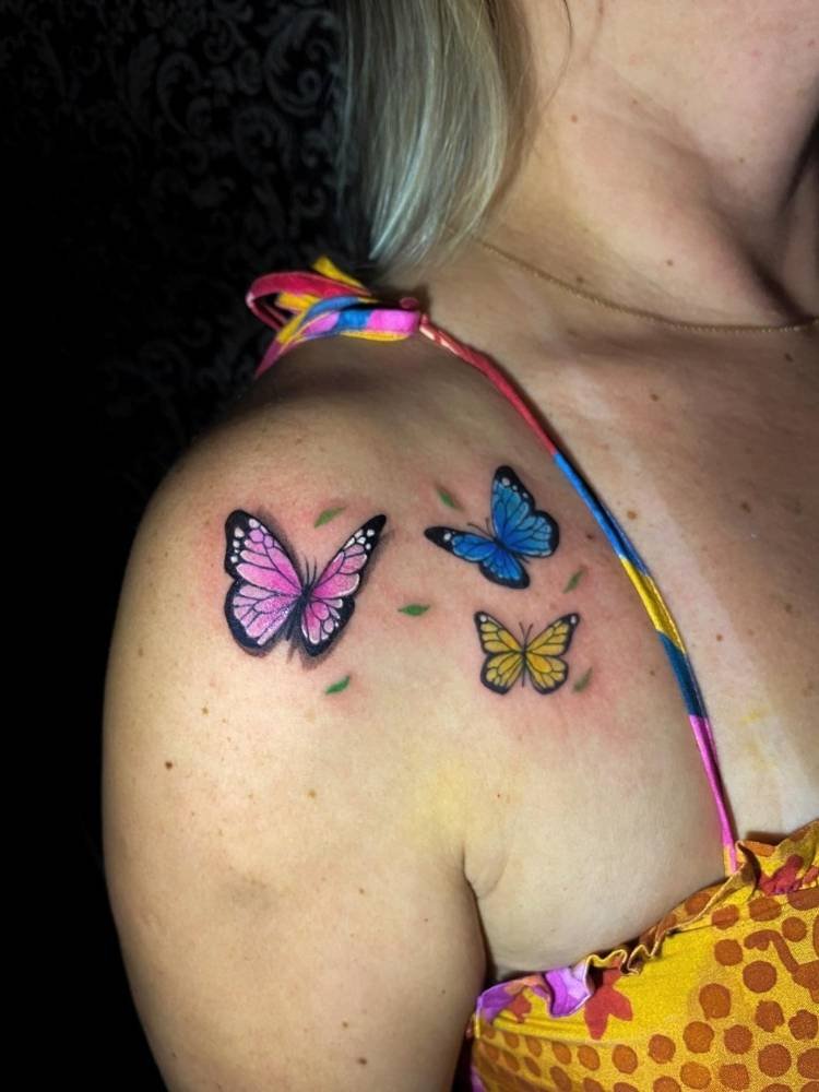 coloridas-rosa-azul-amarelo-borboleta Mulher loira de pele clara com tatuagem de três borboletas coloridas no ombro, uma azul, outra roxa e outra amarela.