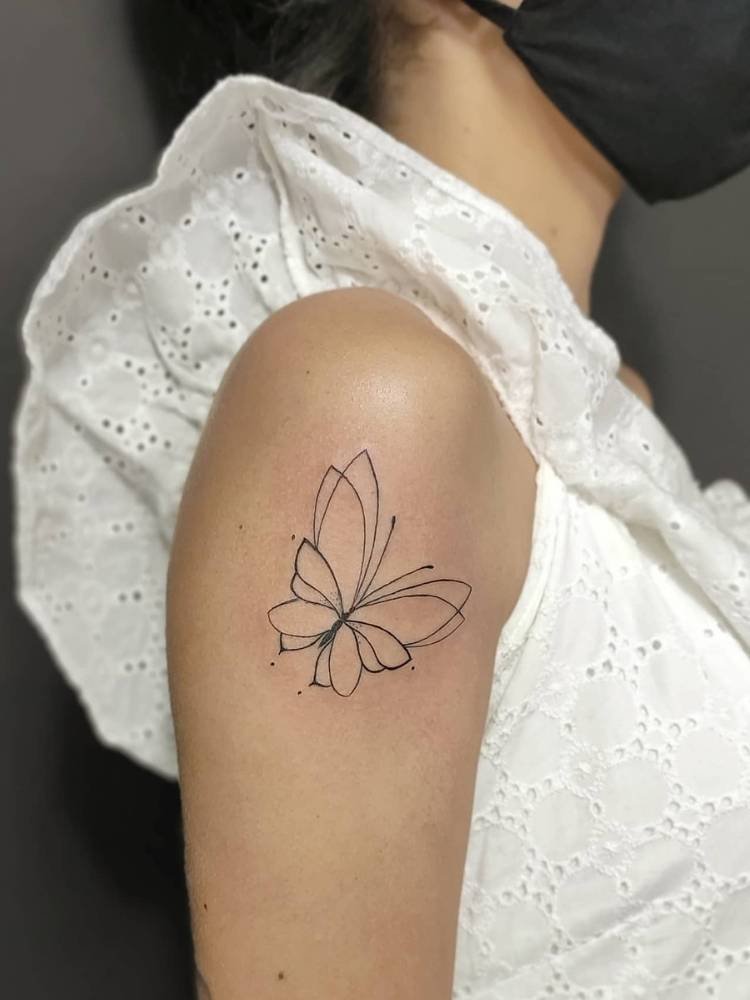 delicada-traco-fino-borboleta Tatuagem de borboleta com poucos traços, sendo eles finos e poucos detalhes. Feita no braço, próxima ao ombro, de uma mulher de pele clara vestindo uma blusa branca.