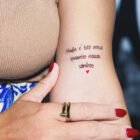 destaque-frases-para-tatuagem-140x140-1-9 A imagem apresenta uma tatuagem delicada no braço de uma mulher com a frase: "Nada é tão nosso quanto nossos sonhos". Abaixo da frase, há um pequeno coração vermelho. Detalhes incluem unhas pintadas e acessórios elegantes.