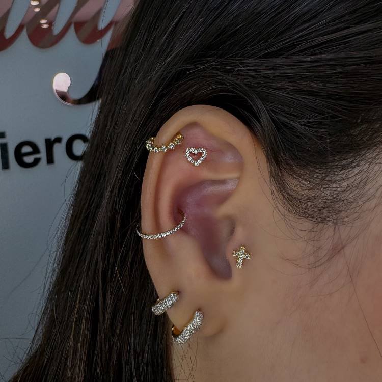 Orelha com piercings delicados. como duas argolas no lóbulo, uma argola na hélix, um coração na scapha e uma cruz no tragus.