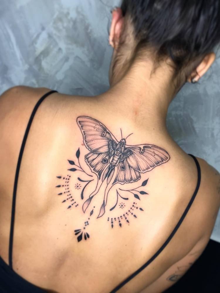 grande-costas-borboleta Tatuagem grande e chamativa de uma borboleta com traços grossos, sombreada, ramos e arabescos. nas costas de uma mulher de pele morena e cabelo preso.