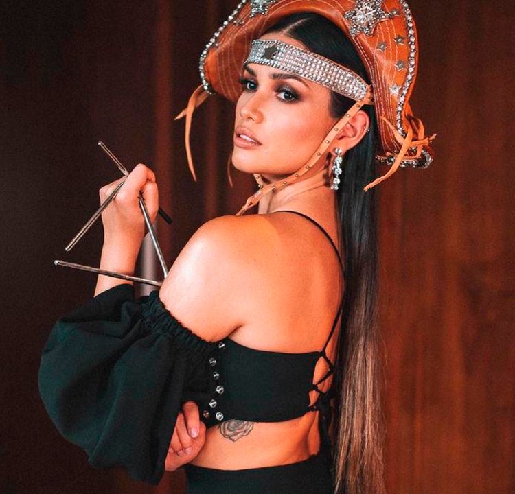 Juliette Freire tatuagem nova Foto de Juliette Freire de lado, com calça e top pretos e chapéu de cangaceiro, segurando o instrumento triângulo. Dá para ver uma parte da tatuagem dela nas costelas.