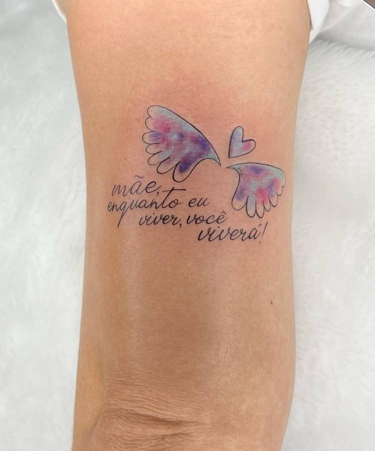 Uma tatuagem delicada no antebraço de uma pessoa. Inclui um par de asas de anjo com tons suaves de roxo e rosa, um pequeno coração e a frase "mãe, enquanto eu viver, você viverá!" em caligrafia.