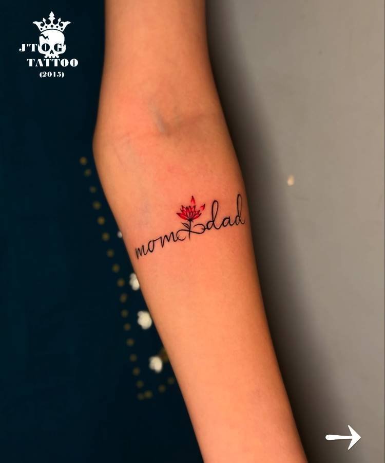 mae-pai-tatuagem-11 A imagem mostra uma tatuagem no antebraço de uma pessoa com as palavras "mom & dad" em uma fonte cursiva preta, onde o "e" de "mom" é substituído por uma flor de lótus vermelha. O fundo é cinza escuro. No canto superior esquerdo há um logotipo e um texto em branco.