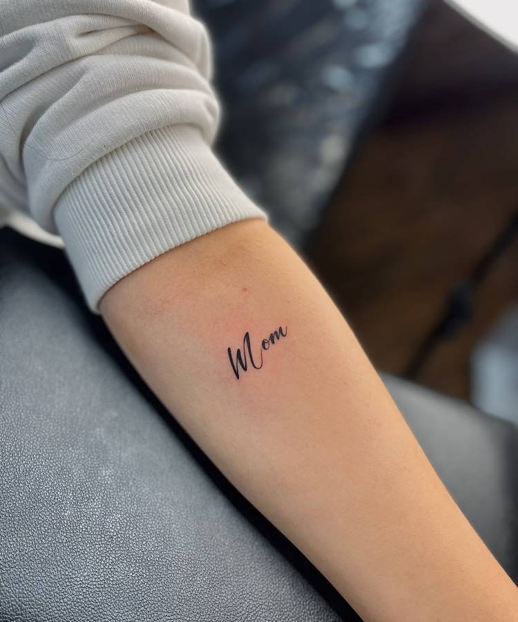 Uma tatuagem delicada e minimalista da palavra "Mom" em caligrafia preta está visível no antebraço de uma pessoa, que veste uma manga comprida branca. A tatuagem se destaca na pele clara, com o braço apoiado em uma superfície escura.