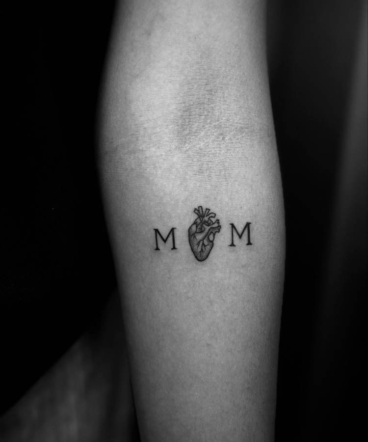 mae-pai-tatuagem-17 A imagem em preto e branco mostra uma tatuagem minimalista no antebraço de uma pessoa. A tatuagem consiste nas letras "M M" com um coração anatômico detalhado entre elas, formando a palavra "MOM". A pele ao redor é clara e lisa.