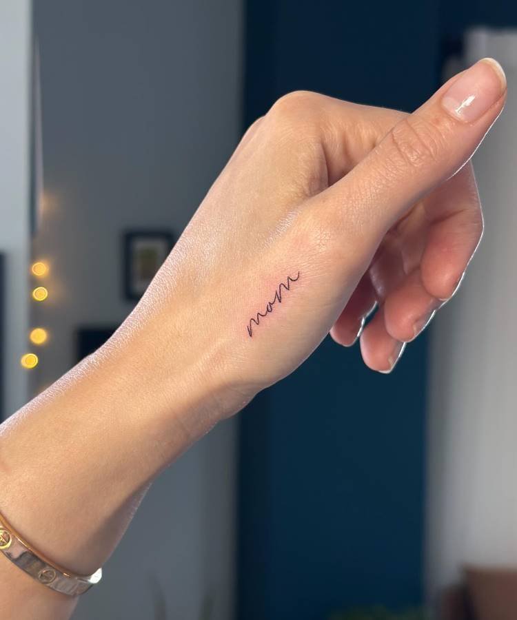 mae-pai-tatuagem-18 A imagem mostra a parte superior da mão de uma pessoa, com uma pequena tatuagem minimalista e fina da palavra "mom" escrita em cursiva preta, posicionada ao longo da linha do dedo mindinho. A pessoa usa uma pulseira dourada. O fundo está desfocado.