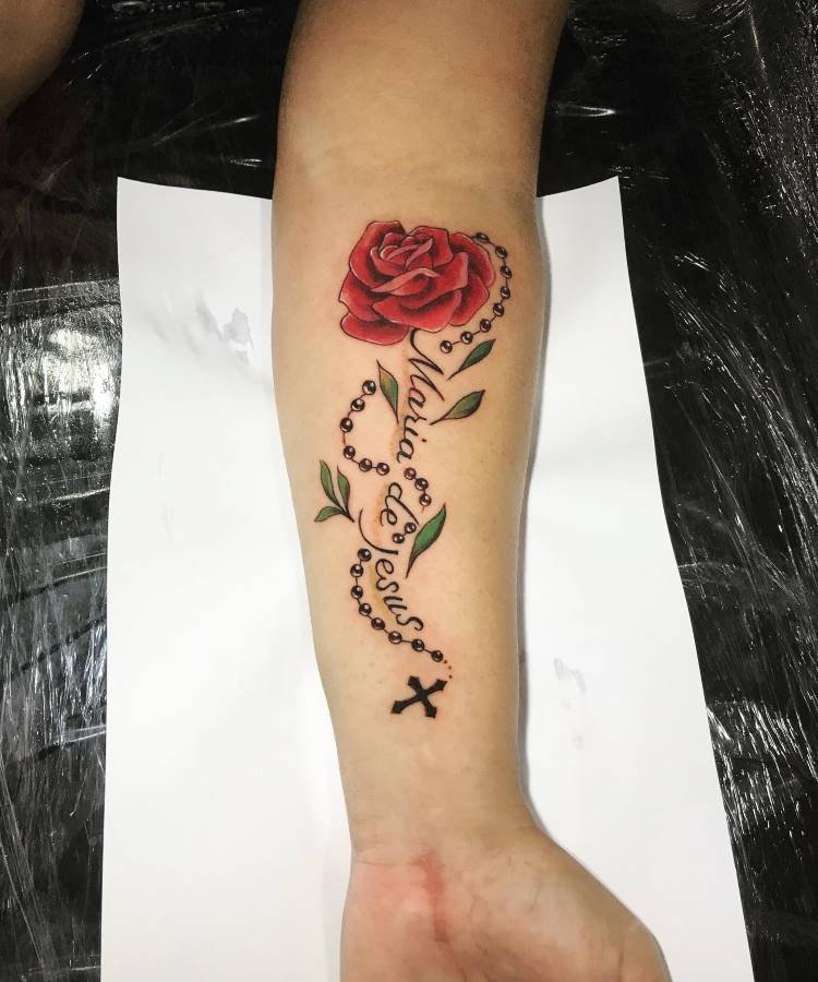 Uma tatuagem vibrante no antebraço, com uma rosa vermelha desabrochando no topo. Um rosário com folhas verdes e a escrita "Maria de Jesus" envolve a rosa, terminando em uma cruz na parte inferior do braço.