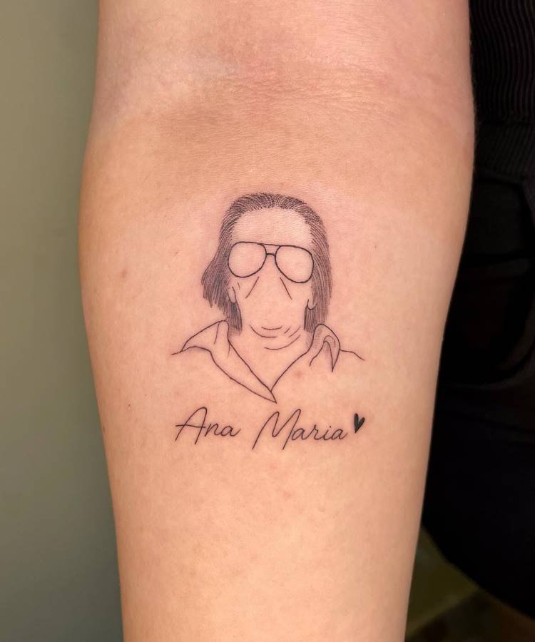 Uma tatuagem minimalista de linha fina no antebraço esquerdo de uma pessoa retrata um busto de cabelo comprido e óculos, com o rosto sem detalhes faciais. Abaixo, o nome "Ana Maria" é escrito em caligrafia com um pequeno coração.