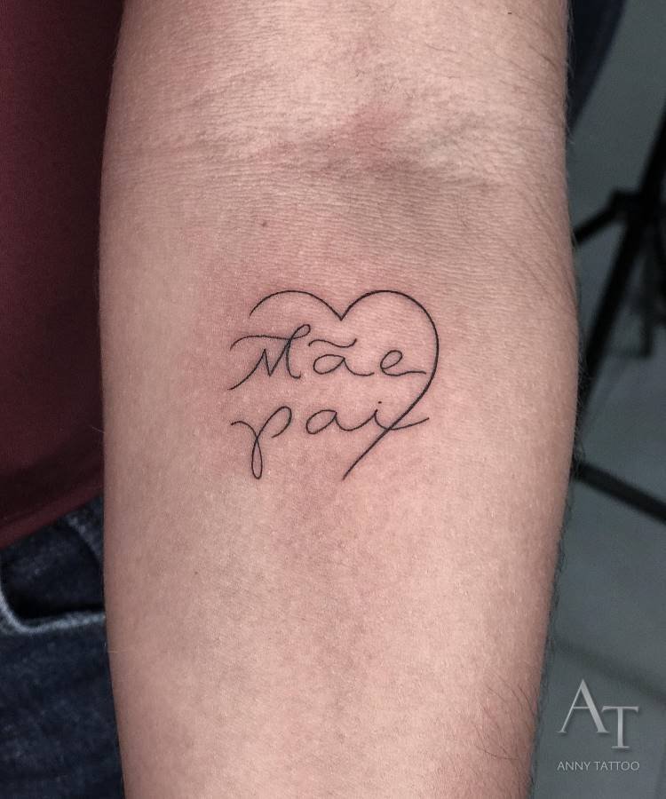 mae-pai-tatuagem-21 A imagem exibe uma tatuagem no antebraço de uma pessoa, consistindo no contorno de um coração com as palavras "Mãe" e "Pai" escritas em caligrafia cursiva dentro dele. A tatuagem é em preto, e a pele é clara. No canto inferior direito, há uma assinatura digital "AT ANNY TATTOO".