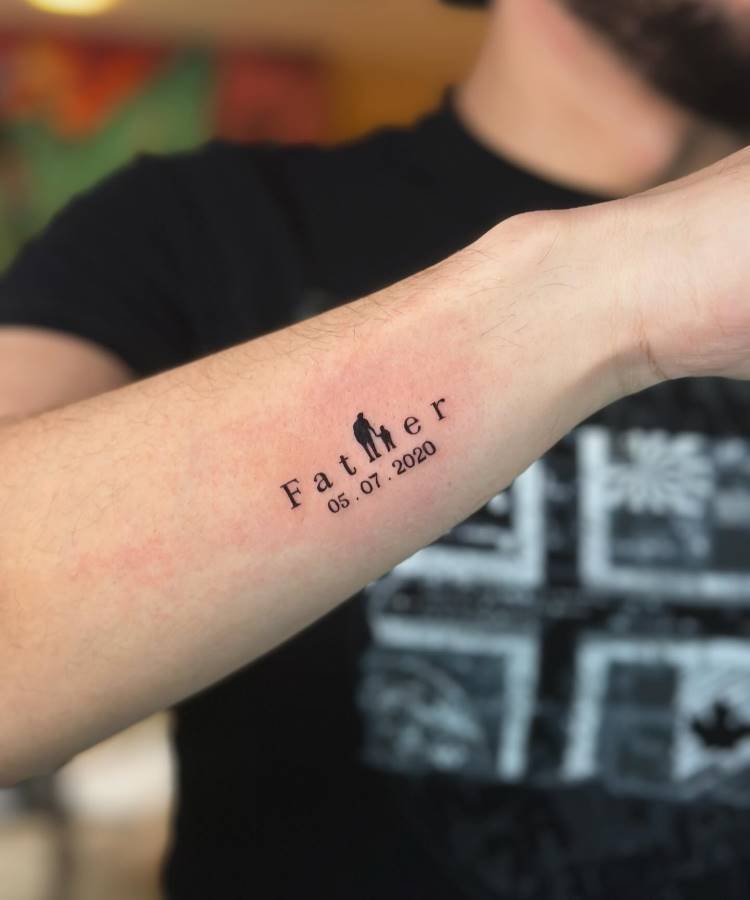 Uma tatuagem minimalista no antebraço de um homem exibe a palavra "Father", onde a letra "h" é substituída pela silhueta de um homem segurando a mão de uma criança. Abaixo, a data "05.07.2020" está marcada, provavelmente representando um nascimento.