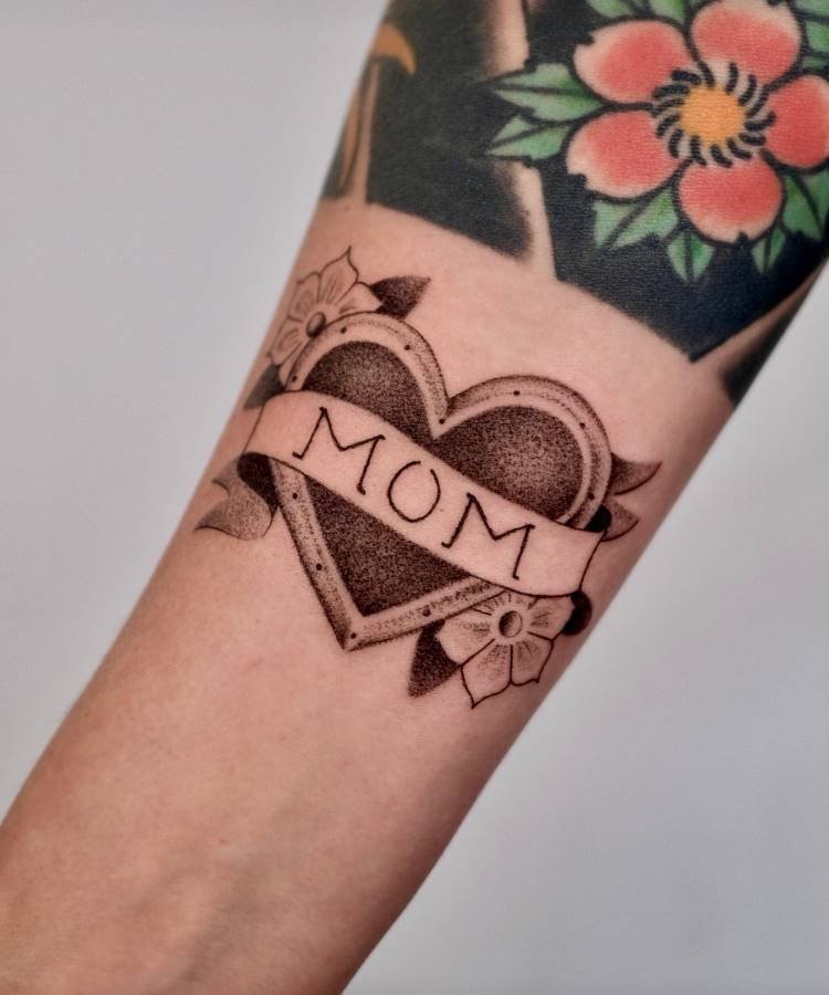 Uma tatuagem de coração em estilo tradicional com o contorno em negrito e preenchimento pontilhado está no antebraço. Uma faixa com a palavra "MOM" envolve o coração, adornado com flores brancas nas laterais. Outra tatuagem colorida de flores é visível acima.