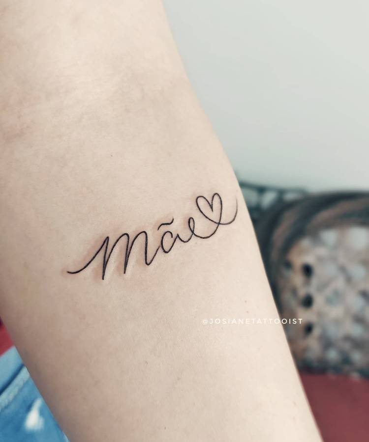Uma tatuagem minimalista e delicada da palavra "Mãe" com um pequeno coração no lugar do til está visível no antebraço de uma pessoa. A fonte é cursiva e fina, e o fundo é um braço claro, com o tecido da roupa levemente visível no canto inferior.