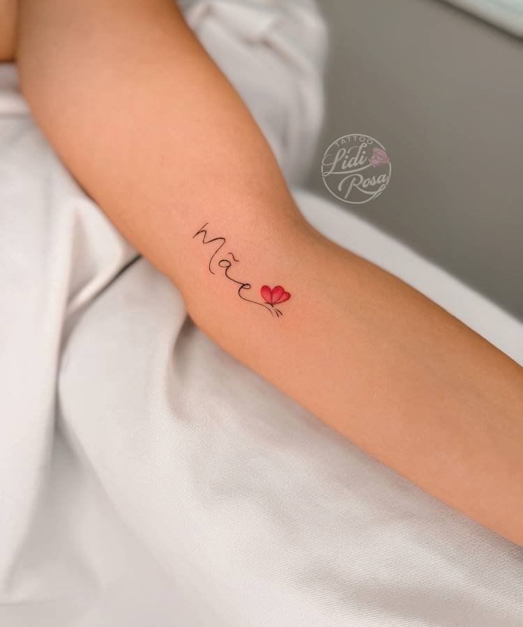 Uma pequena tatuagem da palavra "Mãe" com um coração vermelho substitui a letra "ã" está no antebraço esquerdo de uma pessoa. O desenho é delicado e está posicionado no meio do antebraço.