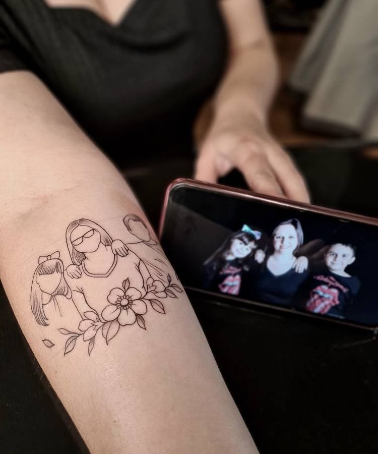 Uma tatuagem de linha fina mostra uma mãe e três crianças abraçadas, com flores e folhas na base. Um celular ao lado exibe a foto original que inspirou o desenho, ilustrando a conexão familiar.