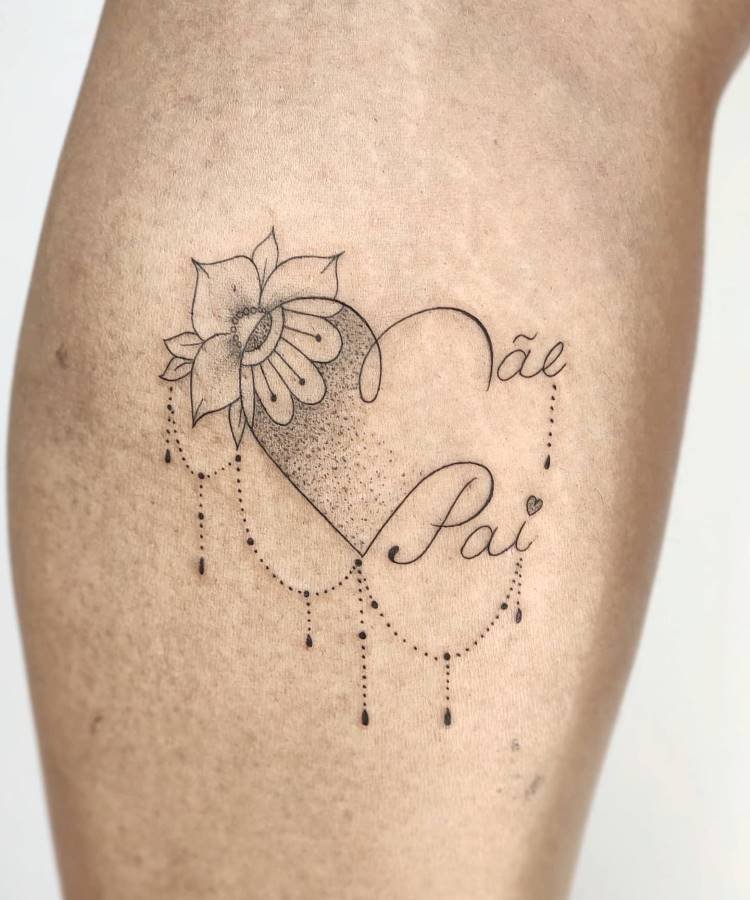 mae-pai-tatuagem-28 A imagem apresenta uma tatuagem detalhada na perna de uma pessoa, com um coração sombreado por pontos. Uma flor delicada brota do topo do coração, enquanto a parte inferior é adornada com linhas pontilhadas e pingentes. As palavras "Mãe" e "Pai" estão escritas em cursiva dentro e ao redor do coração.