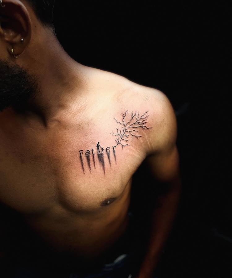 mae-pai-tatuagem-29 A imagem mostra o torso de um homem com uma tatuagem no peito, no lado direito. A tatuagem representa a palavra "Father" com letras que se transformam em galhos de árvores, estendendo-se em direção ao ombro. O fundo da imagem é escuro, criando um contraste com a pele clara.