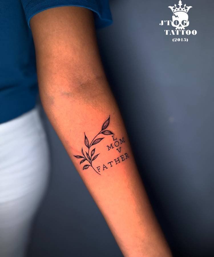 Uma tatuagem delicada no antebraço de uma pessoa. Inclui um ramo com folhas e as palavras "MOM" e "FATHER" dispostas verticalmente ao lado. A palavra "LOVE" também é formada pelas iniciais das palavras "MOM" e "FATHER", criando uma homenagem criativa.