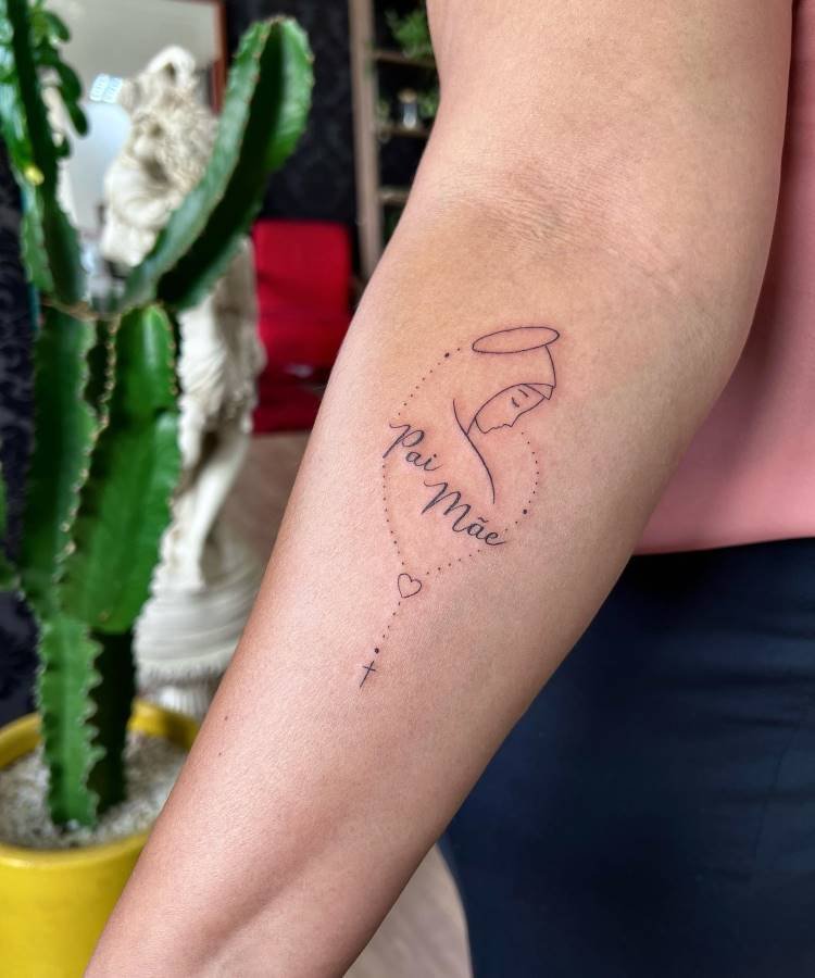 mae-pai-tatuagem-30 A imagem exibe uma tatuagem no antebraço de uma pessoa, apresentando o contorno de uma figura religiosa com auréola, cercada por uma linha pontilhada em forma de coração. As palavras "Pai Mãe" estão escritas em cursiva. Uma cruz e um pequeno coração finalizam o desenho. Um cacto é visível no fundo.