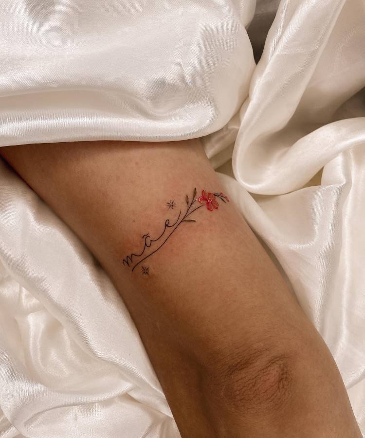 Uma delicada tatuagem da palavra "mãe" com um ramo de flores vermelhas e pequenos brilhos está no antebraço de uma pessoa. O braço repousa sobre um tecido branco amassado, realçando o design minimalista da tatuagem.